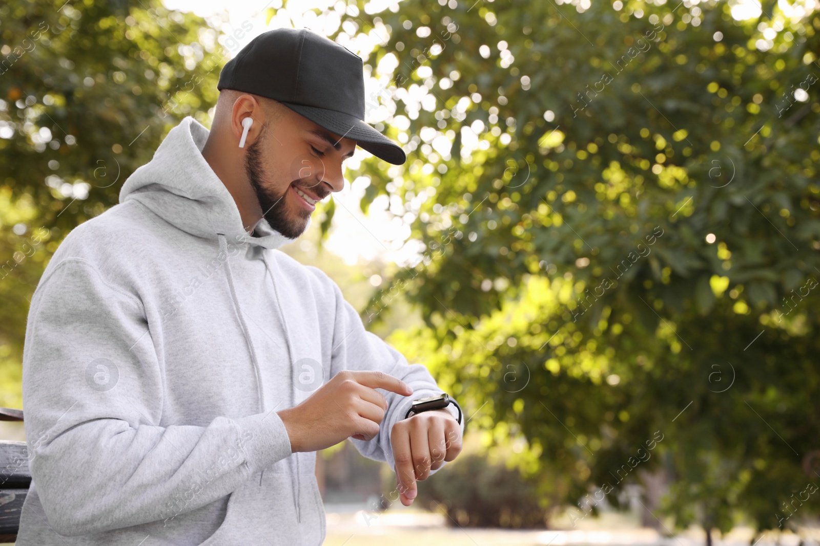 Photo of Young man with wireless headphones and smartwatch listening to music in park. Space for text