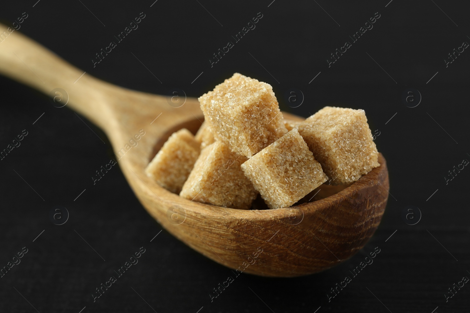 Photo of Brown sugar cubes in spoon on black table, closeup