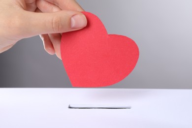 Woman putting red heart into slot of donation box against grey background, closeup