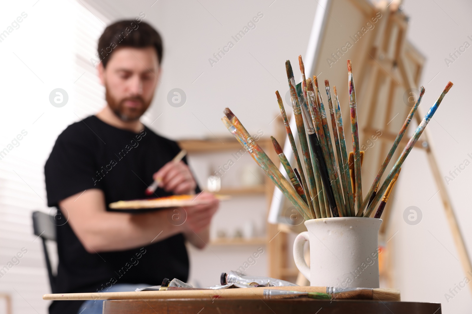 Photo of Man painting in studio, focus on holder with brushes. Using easel to hold canvas