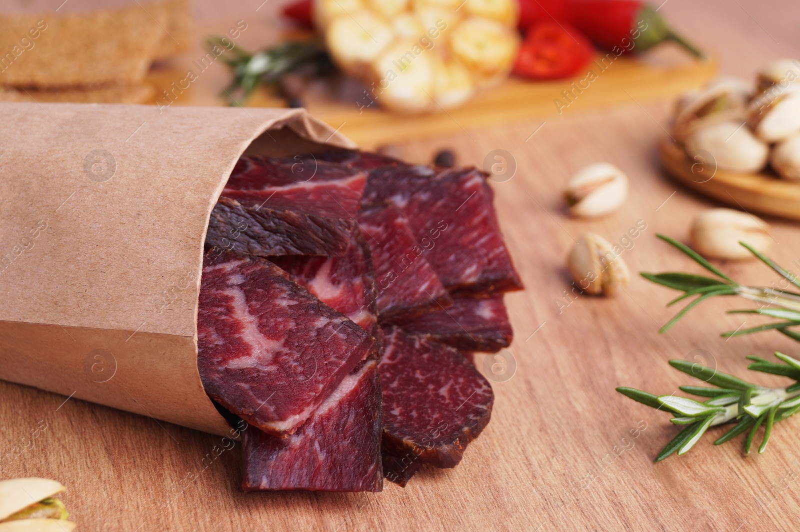 Photo of Paper bag with pieces of delicious beef jerky and different spices on wooden table, closeup