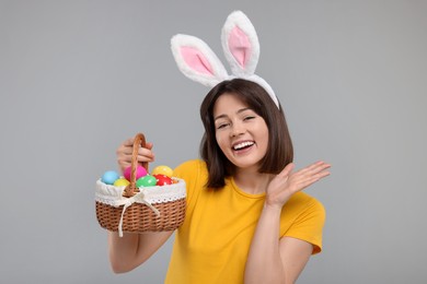 Photo of Easter celebration. Happy woman with bunny ears and wicker basket full of painted eggs on grey background