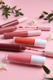 Photo of Different lip glosses, applicators, flowers and green leaves on pink background