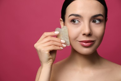 Beautiful young woman doing facial massage with gua sha tool on pink background, closeup