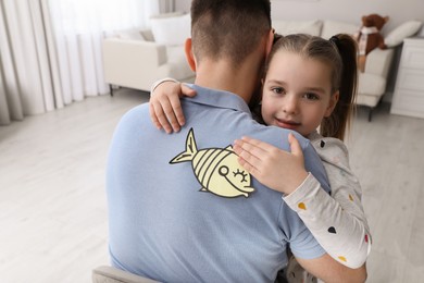Photo of Cute little girl sticking paper fish to father's back at home