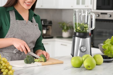 Photo of Young woman cutting broccoli for smoothie at white table in kitchen, closeup