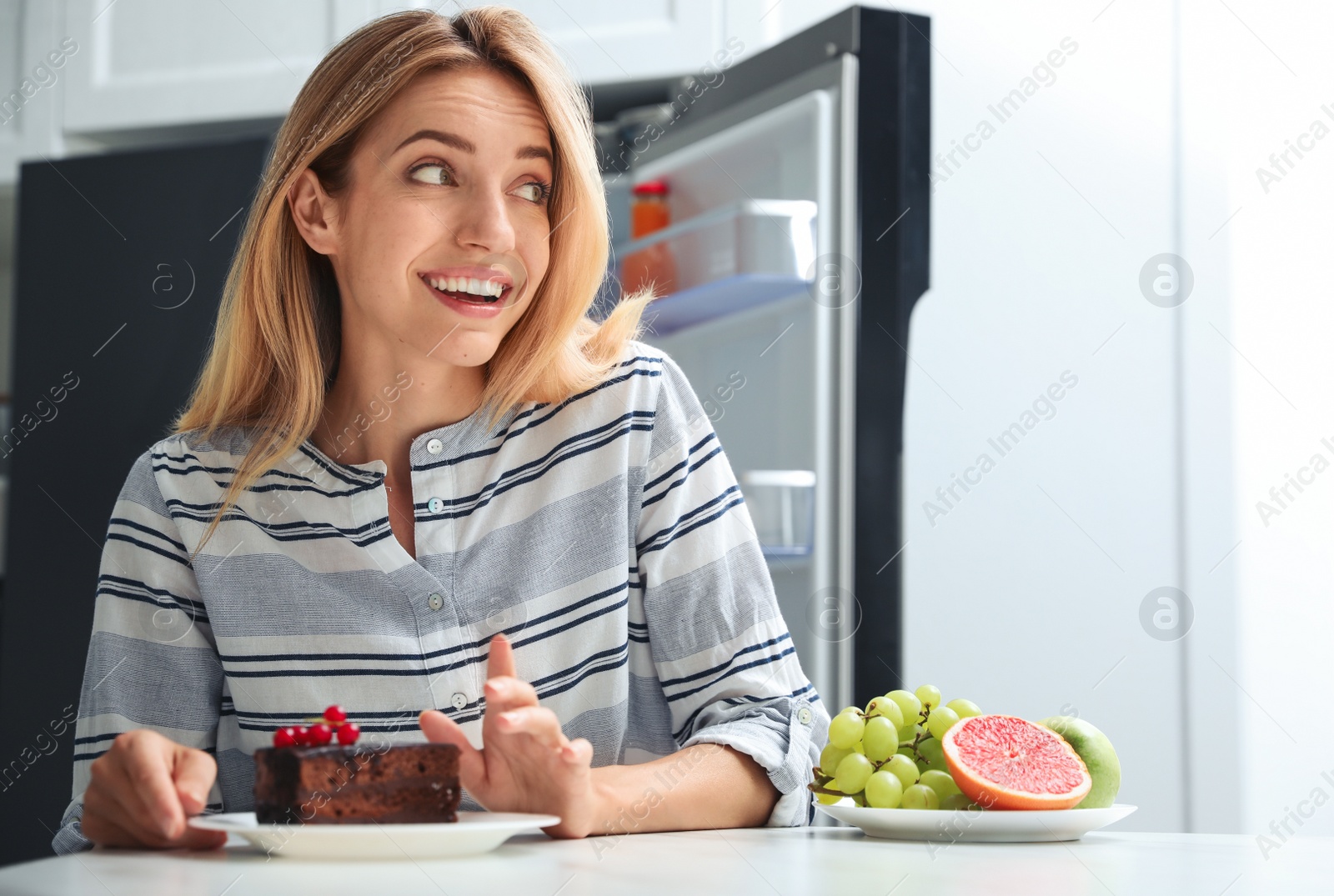 Photo of Woman choosing between cake and healthy fruits at table in kitchen