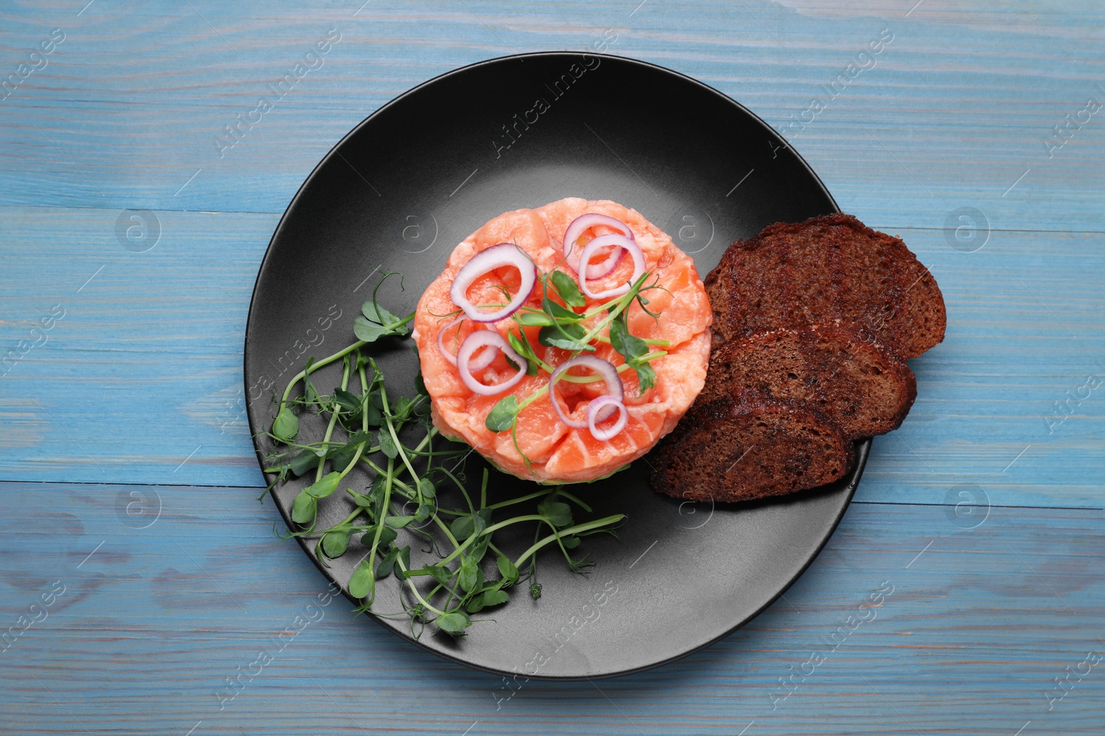 Photo of Tasty salmon tartare with croutons, microgreens and red onion on light blue wooden table, top view