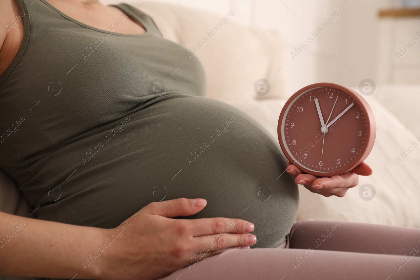 Photo of Young pregnant woman holding clock near her belly at home, closeup. Time to give birth