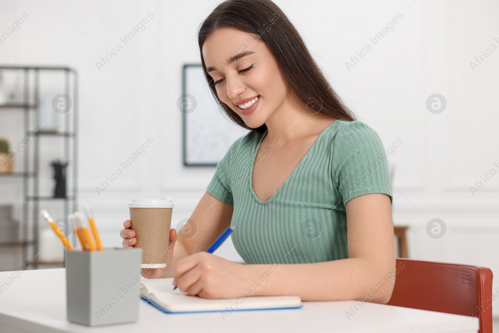 Photo of Young woman with cup of coffee writing in notebook at white table indoors