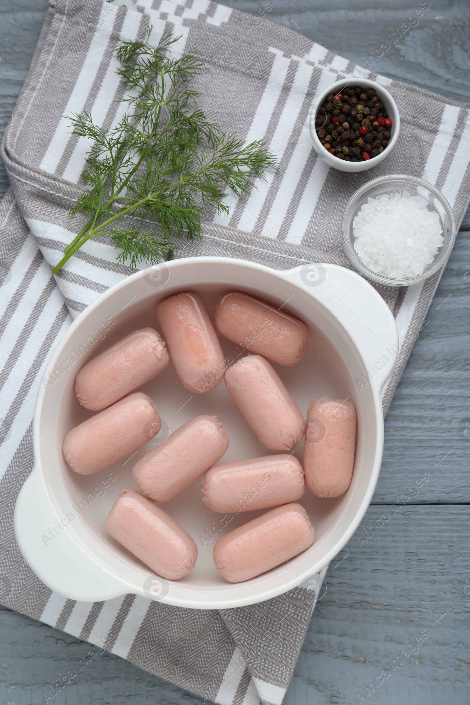 Photo of Bowl of delicious sausages, dill, salt and pepper on grey wooden table, flat lay