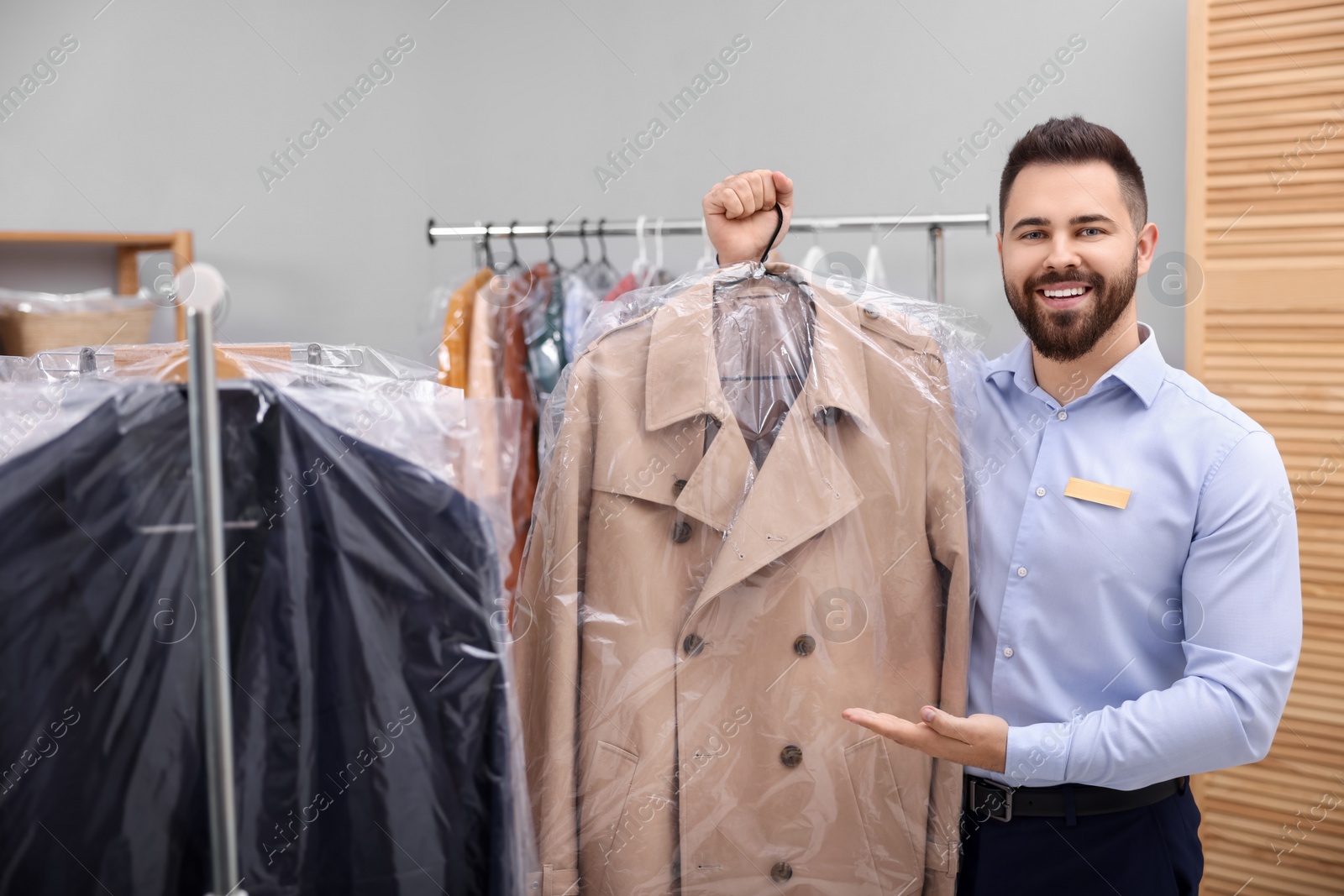 Photo of Dry-cleaning service. Happy worker holding hanger with coat in plastic bag indoors
