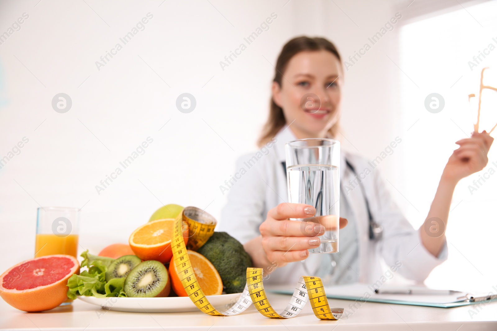 Photo of Nutritionist with glass of water, fruits, vegetables and measuring tape in office