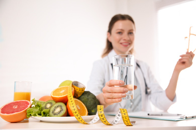 Photo of Nutritionist with glass of water, fruits, vegetables and measuring tape in office