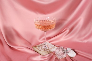 Glass of alcohol drink, perfume and jewelry on pink cloth