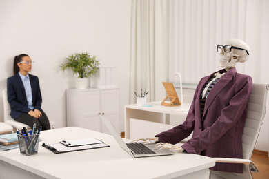 Photo of Human skeleton in suit using laptop at table and woman waiting on chair in office