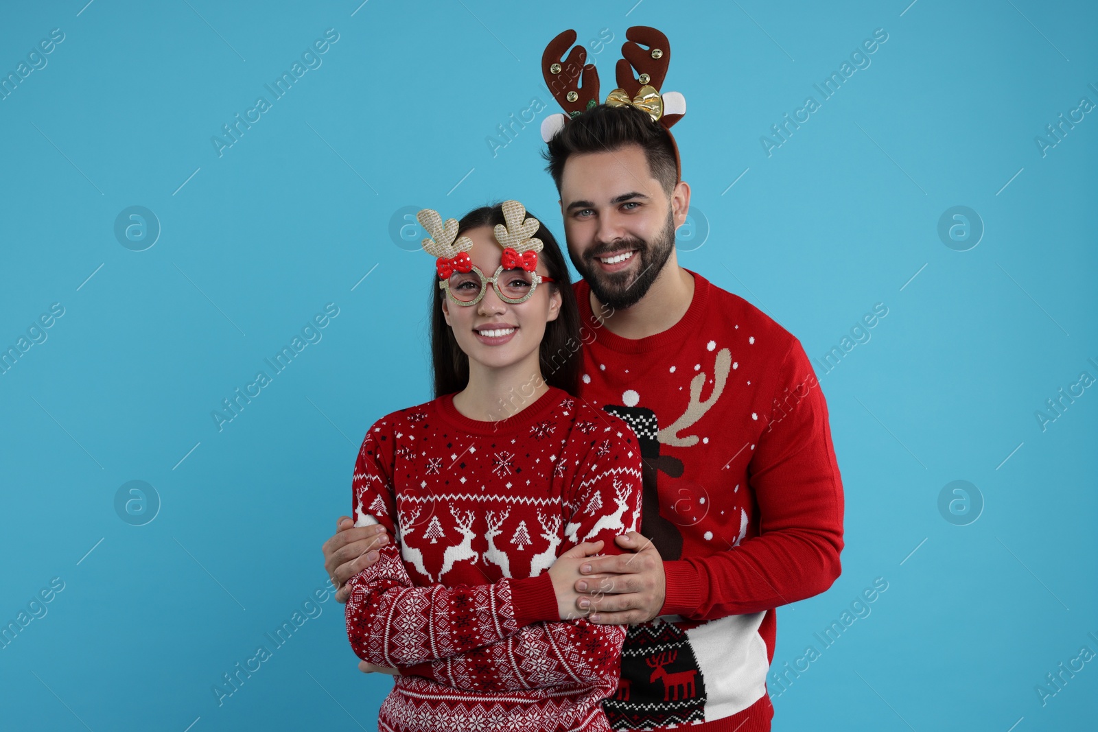 Photo of Happy young couple in Christmas sweaters, reindeer headband and funny glasses on light blue background