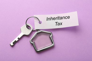 Photo of Card with phrase Inheritance Tax attached to key with house shaped key chain on violet background, top view