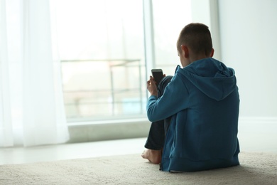 Photo of Upset boy with smartphone sitting near window indoors. Space for text