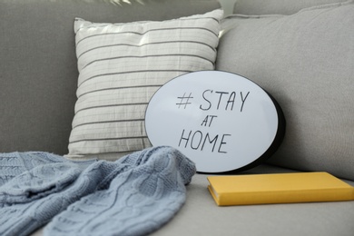 Photo of Book, sweater and speech bubble with hashtag STAY AT HOME on sofa, closeup. Message to promote self-isolation during COVID‑19 pandemic