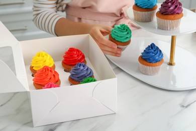 Woman putting delicious colorful cupcakes on dessert stand at white marble table indoors, closeup