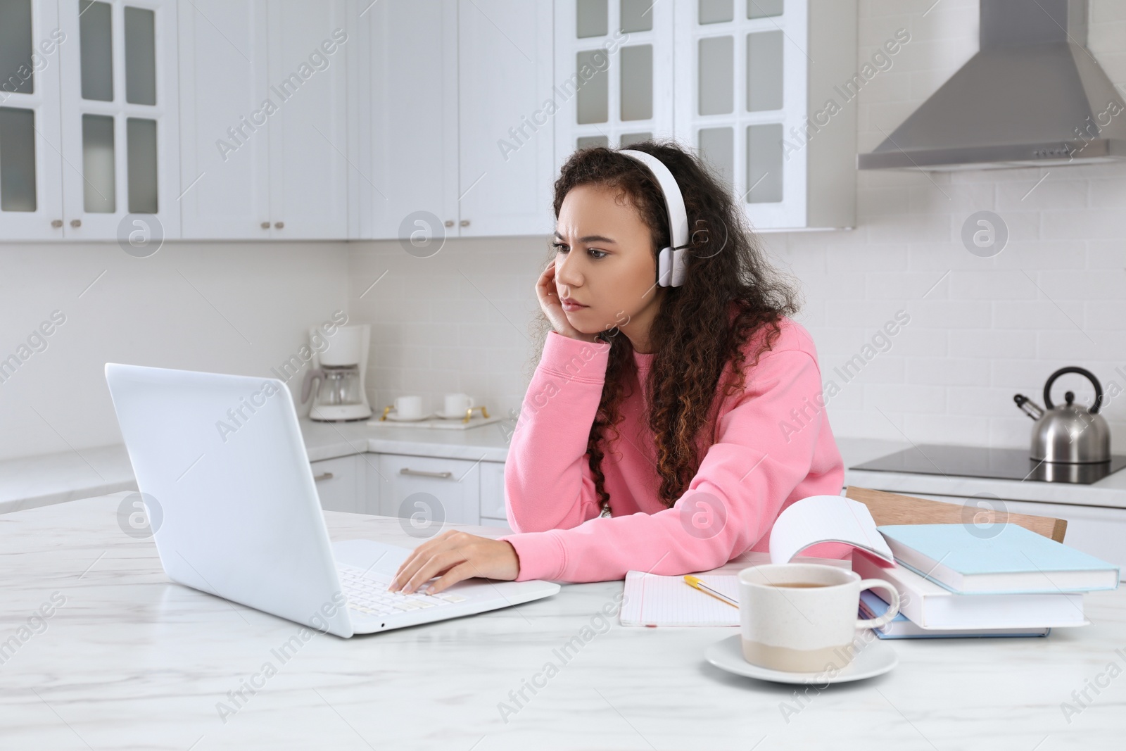 Photo of African American woman with modern laptop and headphones studying in kitchen. Distance learning