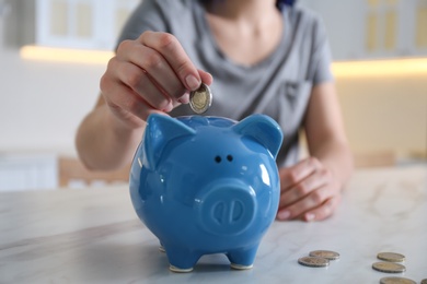 Photo of Woman putting money into piggy bank at marble table indoors, closeup