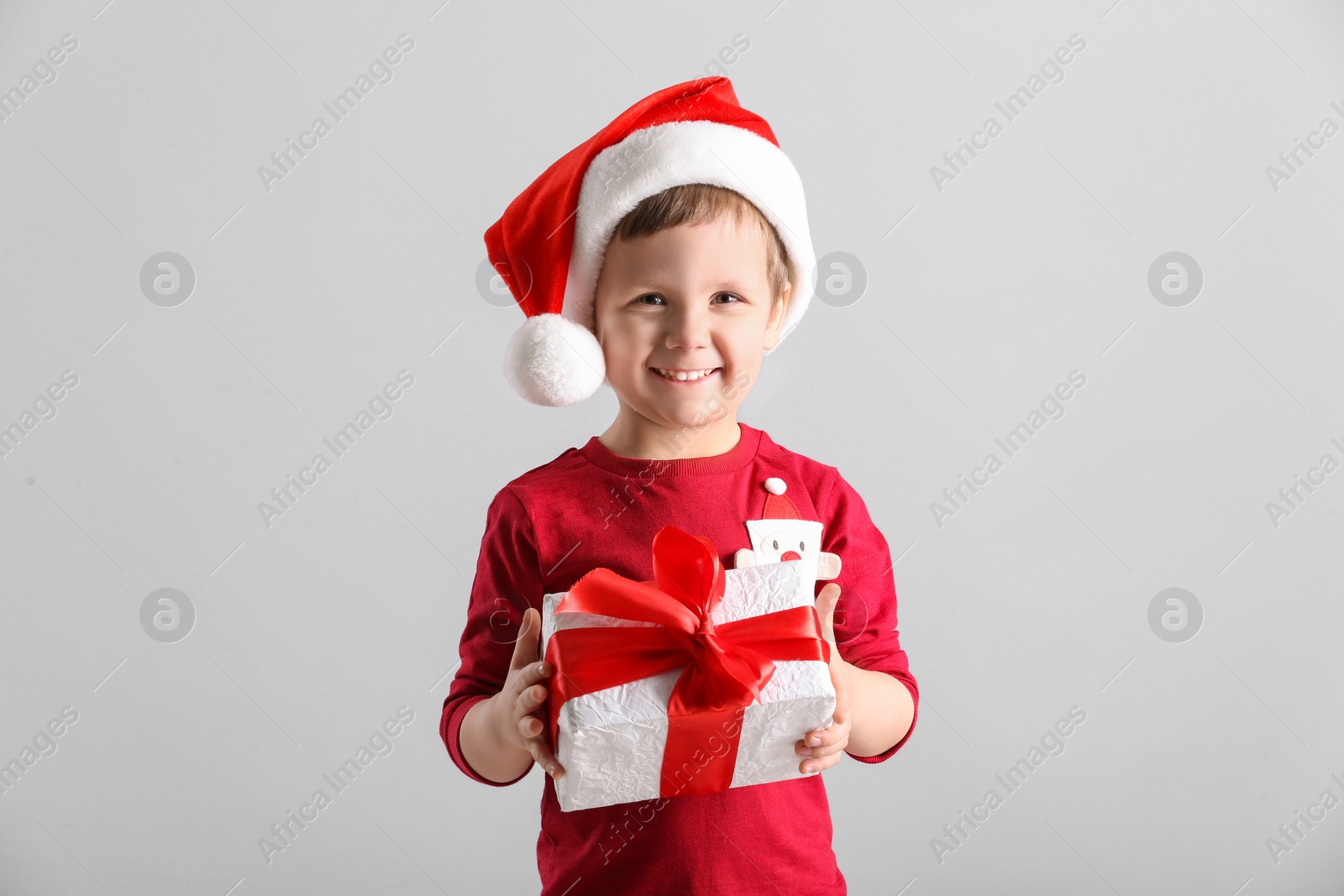 Photo of Cute little boy in Santa Claus hat holding gift box on light grey background