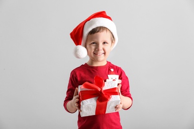 Photo of Cute little boy in Santa Claus hat holding gift box on light grey background