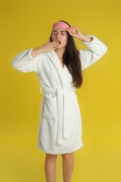 Photo of Beautiful young woman in bathrobe and eye sleeping mask yawning on yellow background