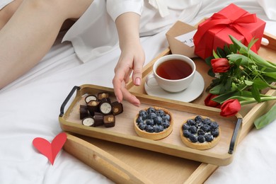 Photo of Tasty breakfast served in bed. Woman with desserts, tea, gift box and flowers at home, closeup