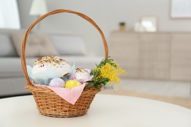 Photo of Basket with delicious Easter cakes, dyed eggs and flowers on white table indoors. Space for text