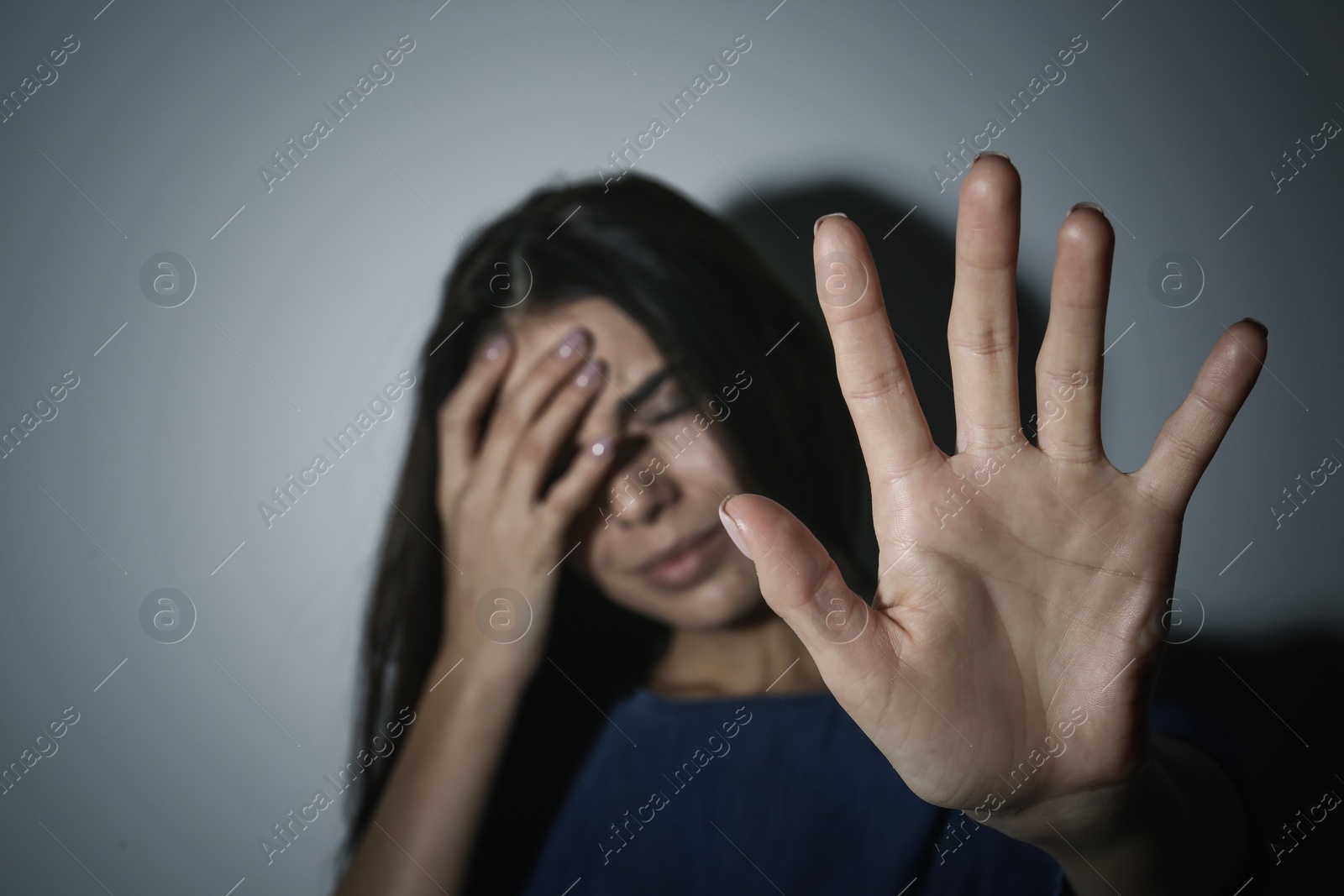 Photo of Crying young woman making stop gesture near white wall, focus on hand. Domestic violence concept