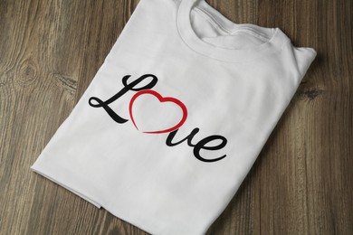 Photo of White shirt with printed word Love and heart shape on wooden table, top view