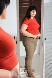 Photo of Overweight woman trying to button up tight trousers in front of mirror at home