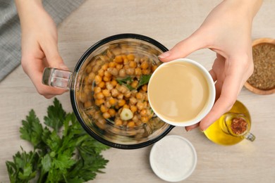 Photo of Woman adding tahina into blender with ingredients for hummus at table, top view