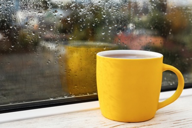 Yellow cup of hot tea on white wooden window sill, closeup. Rainy weather