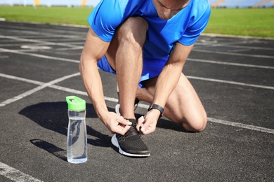 Sporty man tying shoelaces near bottle of water at stadium on sunny day