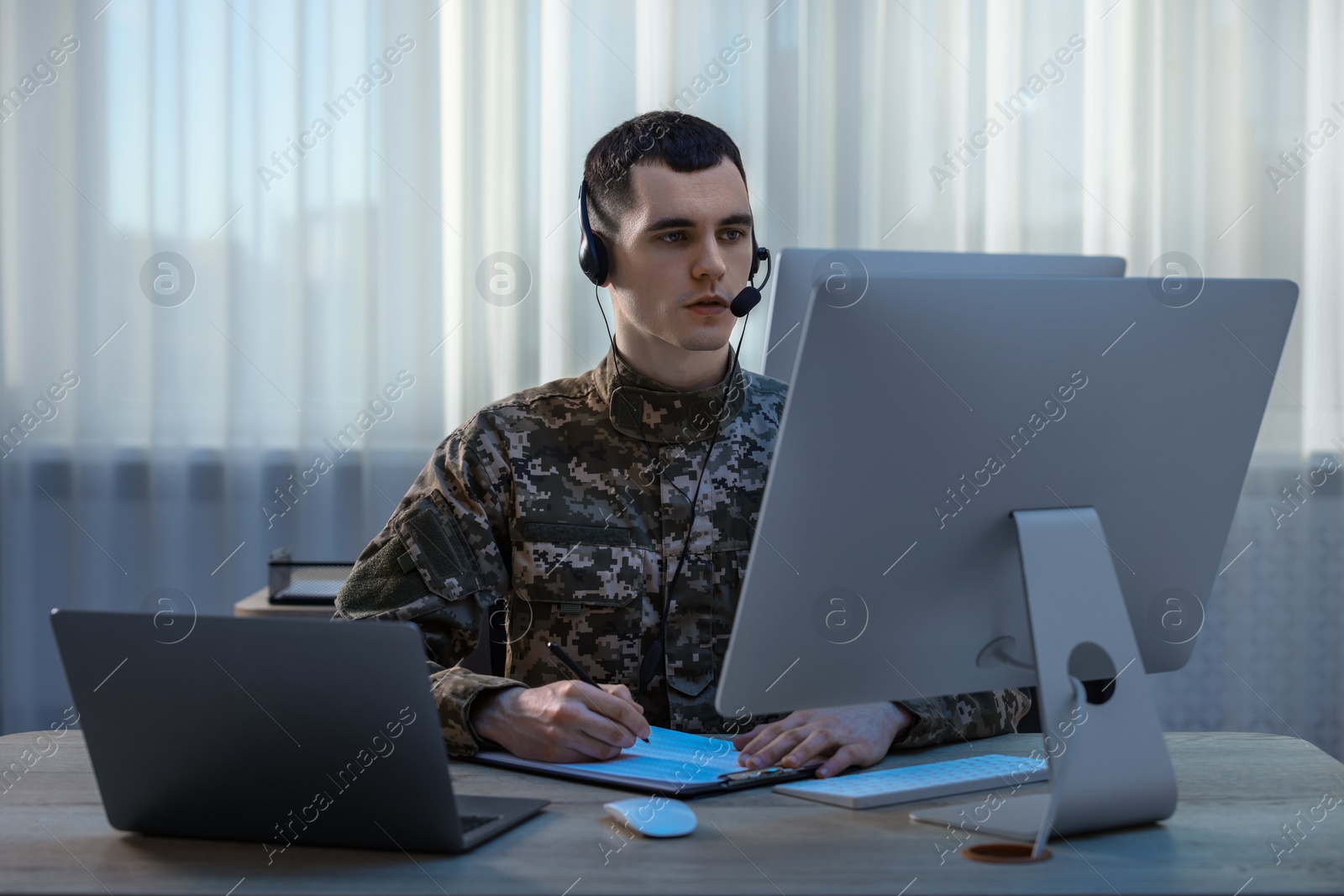 Photo of Military service. Young soldier in headphones working at wooden table in office