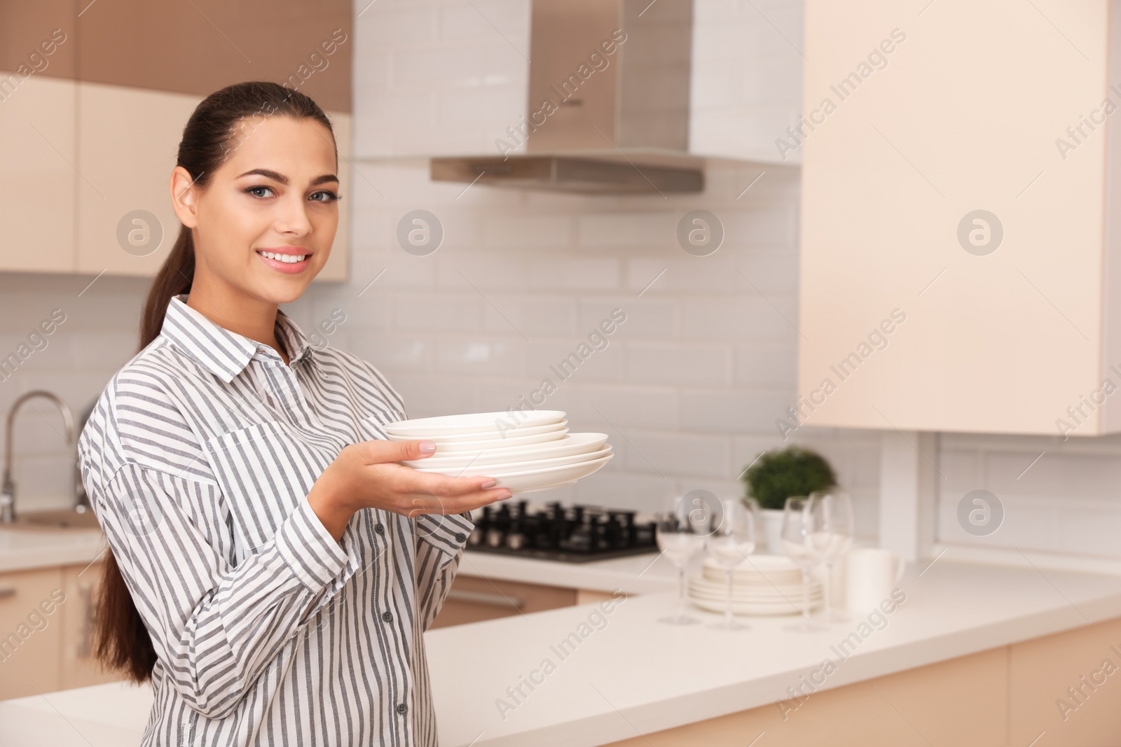 Photo of Beautiful young woman holding stack of clean dishes in kitchen