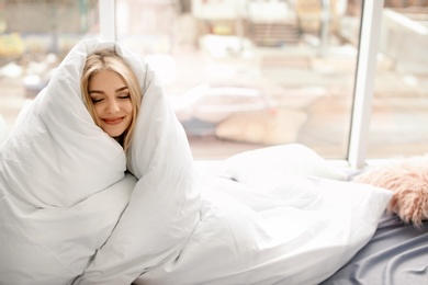 Beautiful young woman wrapped in soft blanket sitting near window at home. Winter atmosphere