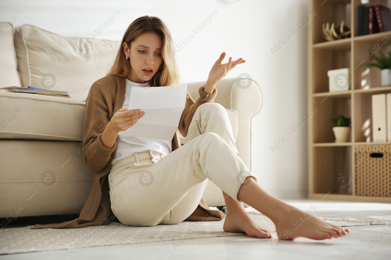 Photo of Shocked woman reading letter while sitting on floor near sofa at home