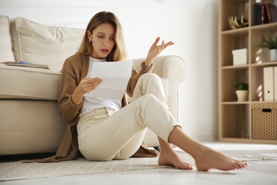 Shocked woman reading letter while sitting on floor near sofa at home