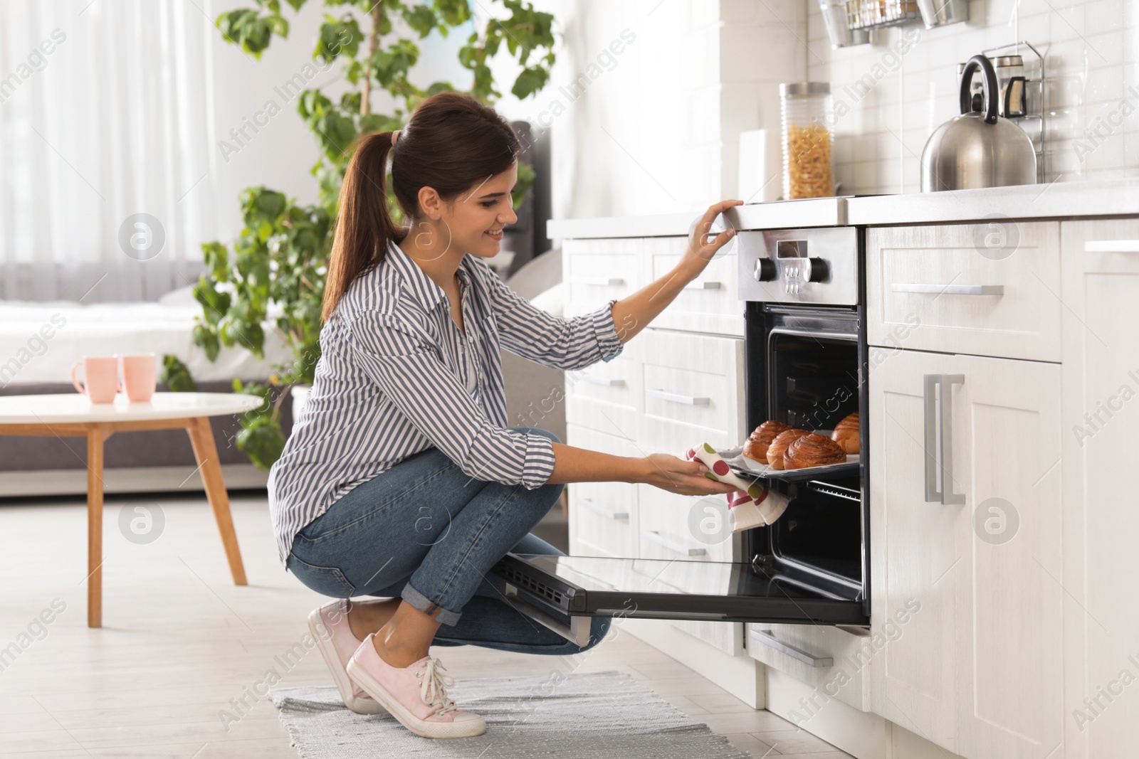 Photo of Beautiful young woman taking out tray of baked buns from oven in kitchen