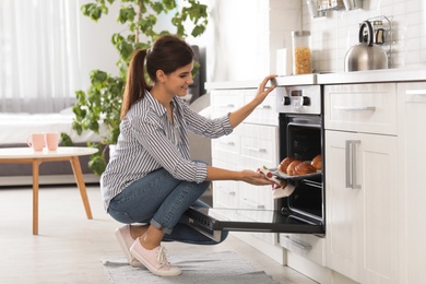 Photo of Beautiful young woman taking out tray of baked buns from oven in kitchen
