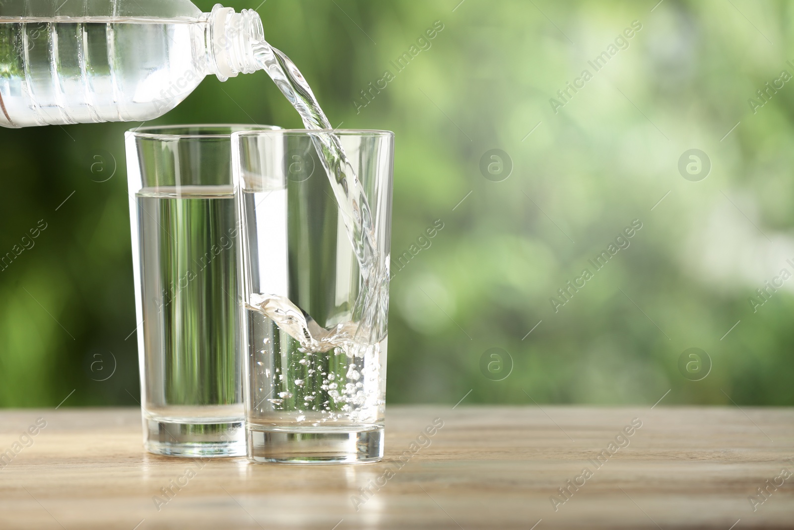Photo of Pouring water from bottle into glass on wooden table outdoors, space for text