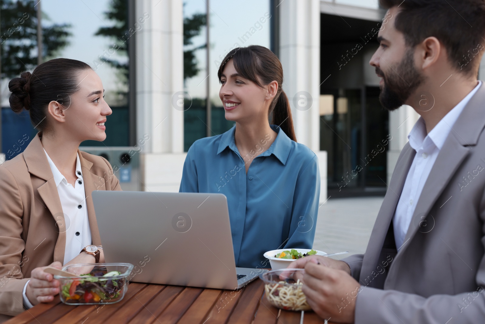 Photo of Business lunch. Happy colleagues with laptop spending time together at wooden table during break outdoors