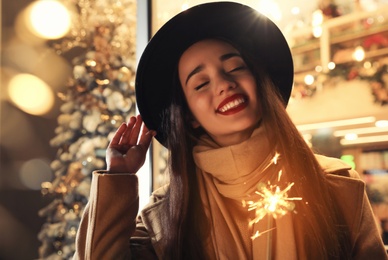 Woman in warm clothes holding burning sparkler on blurred background