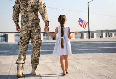 Photo of Soldier and his little daughter with American flag outdoors, back view. Veterans Day in USA
