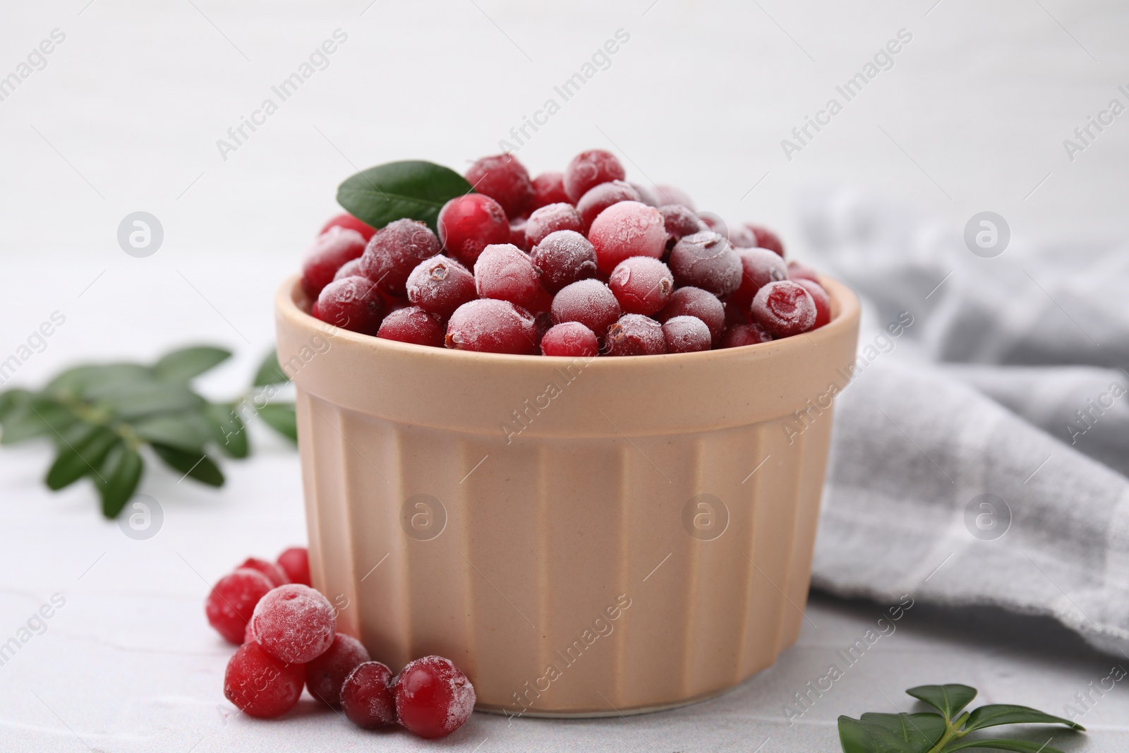 Photo of Frozen red cranberries in bowl and green leaves on white table, closeup